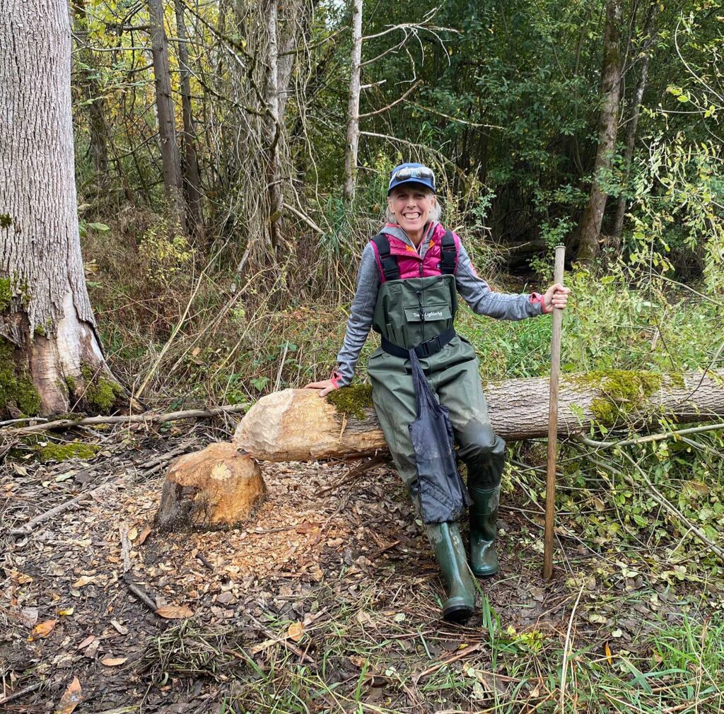 Person sitting on a downed tree in a forest holding stick and dressed in waders, smiles at camera.