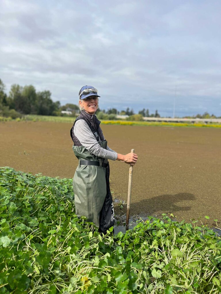 Person in waders standing in bog, holding a walking stick and smiling at the camera