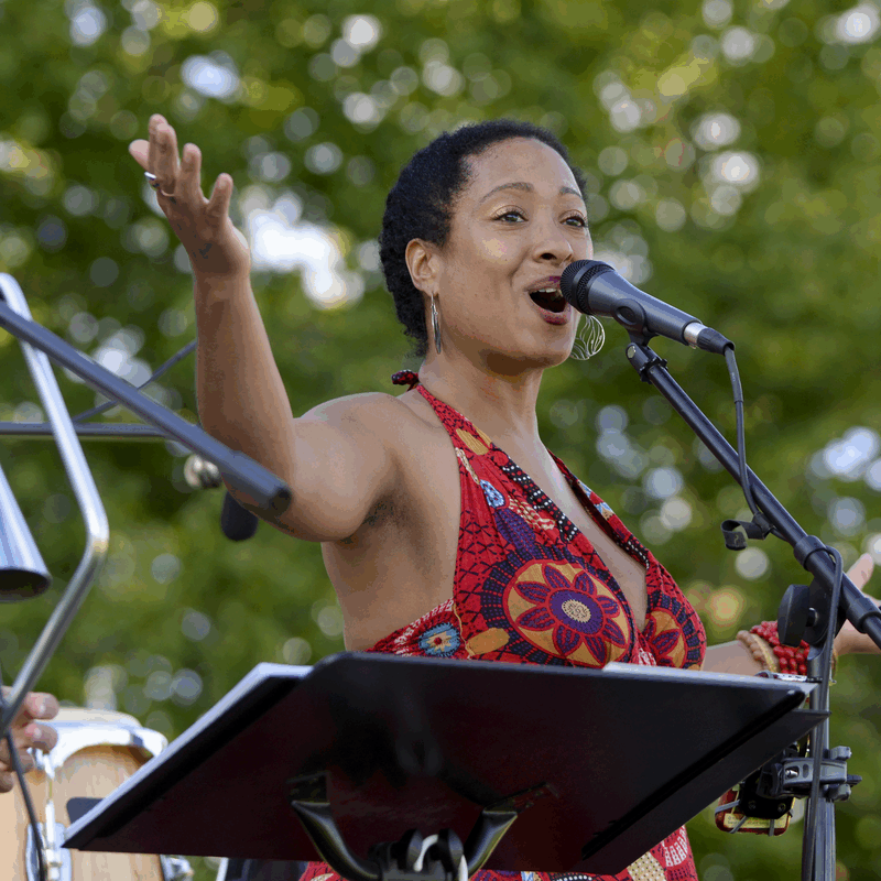 woman standing at a podium singing into a microphone at an outside concert. Other musicians in the background playing instruments.